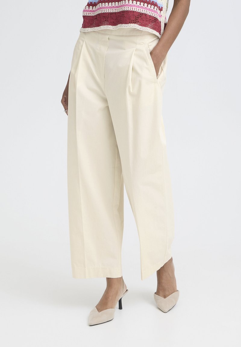 Femme portant un pantalon large crème cropped avec des plis et des talons beiges à bouts pointus, main dans la poche, sur un fond uni.