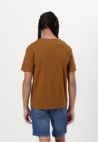 Camiseta de algodón marrón con un ajuste relajado y mangas cortas, vista desde la parte de atrás. Combinada con pantalones cortos de mezclilla azules.