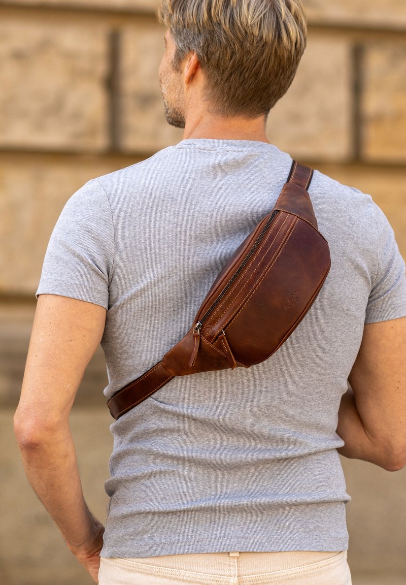 Borsa a tracolla in pelle marrone con una forma curvata, caratterizzata da una chiusura con zip e dettagli cuciti, indossata su una t-shirt grigia su uno sfondo testurizzato.