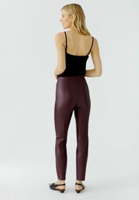 Burgunderfarbene Kunstleder-Leggings mit schmaler Passform, hoher Taille und Nahtdetails. Kombiniert mit einem schwarzen Spaghettiträgertop und schwarzen Schuhen.
