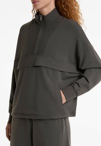 Grauer, strukturierter Pullover mit hohem Kragen und halbem Reißverschluss, der ein geschichtetes Design und Seitentaschen aufweist. Weicher, lässiger Stoff.