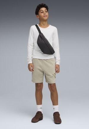 Niño de pie con camiseta blanca de manga larga, pantalones cortos beige, zapatillas marrones, calcetines blancos y bolso cruzado negro sobre fondo gris.