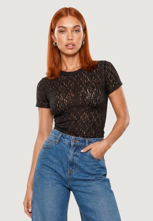 Femme aux cheveux roux portant un haut noir en dentelle à manches courtes et un jean bleu taille haute, posant avec une main dans la poche.
