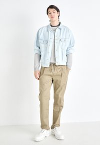 Chaqueta de mezclilla de color azul claro con dos bolsillos en el pecho, suéter gris de canalé debajo, pantalones caqui con cordón y zapatillas deportivas blancas de suela gruesa.