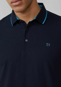 Navy polo shirt made of textured fabric, featuring a contrasting teal collar trim and three-button placket with a small logo on the chest.