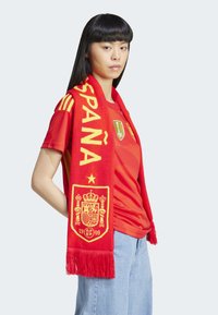 Červený fotbalový šál s žlutým textem "ESPAÑA" a designem královského erbu. Nosený přes odpovídající červený dres; má třásně na koncích a výrazné grafiky.