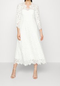 Valkoinen pitsimekko, jossa on kolme neljänneshihaa, istuva vartalonosa ja lippaantuva hame. Ominaisuuksina ovat aaltoileva helma ja herkkä kukkakuvio.