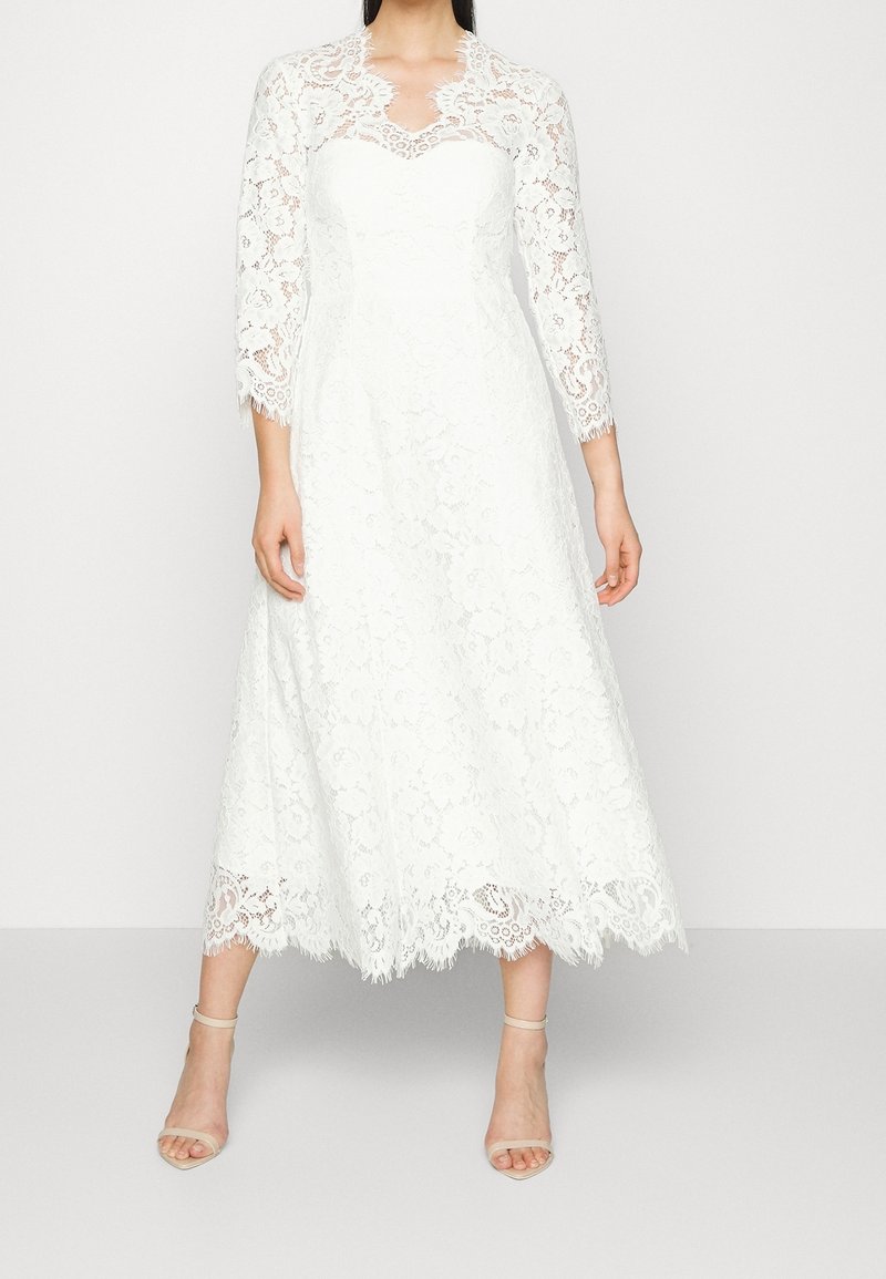 Valkoinen pitsimekko, jossa on kolme neljänneshihaa, istuva vartalonosa ja lippaantuva hame. Ominaisuuksina ovat aaltoileva helma ja herkkä kukkakuvio.