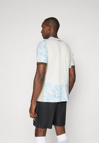 Μπλούζα αθλητικής ομάδας κοντομάνικη σε ανοιχτό μπεζ χρώμα με μπλε μοτίβο paisley στους ώμους και την πλάτη, ελαιόχρωμες λεπτομέρειες, και λογότυπο στον λαιμό.