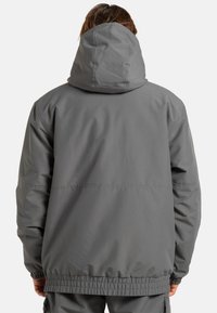 Szara kurtka z kapturem o luźnym kroju, z elastycznymi mankietami i ściągaczem na dole, wykonana z gładkiego, wodoodpornego materiału.