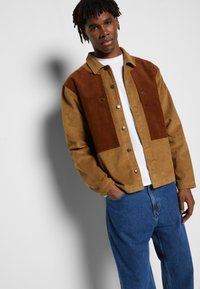 Nuori mies, joka käyttää vaaleanruskeaa ja ruskeaa corduroy-takkia valkoisen paidan päällä ja sinisiä farkkuja, seisoo yksinkertaisen valkoisen seinän edessä varjon kanssa.