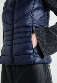 Navy puffer vest with quilted texture, front zipper, and side pockets. Paired with a dark gray wool sleeve detail. Black leather pants visible.