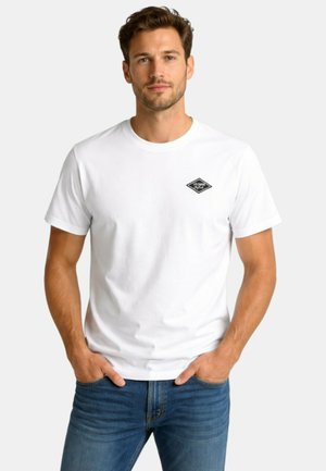 Homme aux cheveux courts portant un t-shirt blanc avec un petit logo noir sur la poitrine et un jean bleu, les mains dans les poches, debout devant un fond uni.