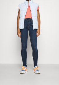 Světle modrá péřová vesta, krátké rukávy, přední zip. V kombinaci s korálovým tričkem a tmavými skinny džíny. Bílé a oranžové tenisky s modrými akcenty.