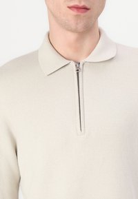 Hele beež pullover, millel on lukuga krae, tekstuuriga kudum ja sirge lõige. Silmapaistev hõbedane luku detail kaelas.