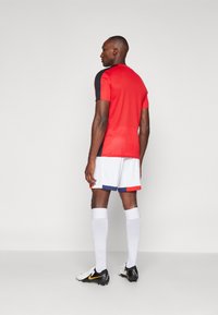 Rode sporttrui met zwarte accenten, witte shorts met blauwe en rode strepen, witte sokken en zwarte voetbalschoenen met gele details.
