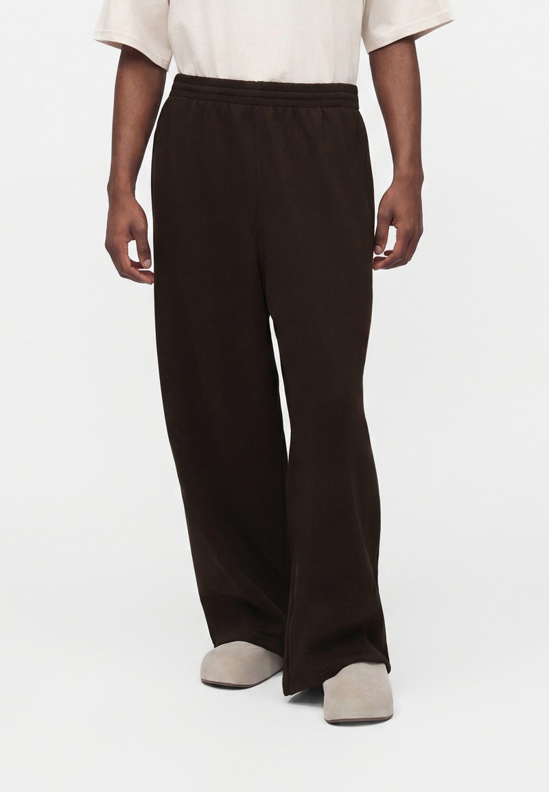 Person iført løse sorte bukser med elastisk talje og beige slip-on sko, stående mod en ensfarvet lys baggrund.