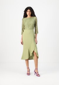 Robe mi-longue en vert clair avec un haut en dentelle et des manches trois-quarts. Présente un ourlet asymétrique à volants et une fente sur le côté.