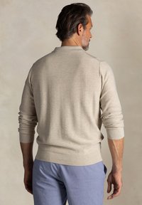 Beige gebreide trui met lange mouwen, ribbelboorden en zoom. Glad textuur, ronde halsontwerp, gedragen met lichtblauwe broek.