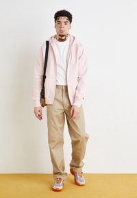 Light pink zip-up hoodie, white shirt underneath, tan trousers, and multicolored sneakers with orange accents; carrying a small brown bag.