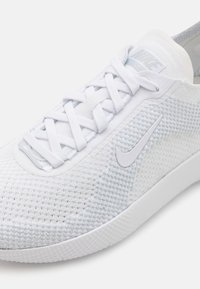 Hvid sportsko med en struktureret mesh-overdel, flade hvide snørebånd, en subtil lys blå detalje og et hvidt Nike-logo på siden.