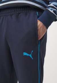 Jūras spēku sporta bikses ar tirkīza sānu svītrām un tirkīza Puma logotipu. Tām ir elastīga josta un sānu kabatas. Mīksta auduma tekstūra.