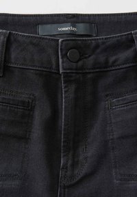 Jeans en denim noir avec poches avant, fermeture à bouton et une étiquette à la taille portant le mot "un jour." en texte blanc.