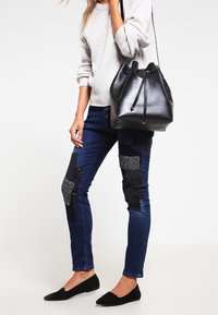 Jeans ajustados de color azul oscuro con acentos de parcheado negro y texturizado, combinados con un suéter gris claro y un bolso tipo cubo de cuero negro.