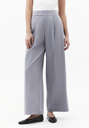 Kobieta ubrana w szare spodnie z wysokim stanem i szerokimi nogawkami, z przodu plisami i detalami z guzikami, w połączeniu z czarnymi balerinami i białym topem, stojąca na białym tle.