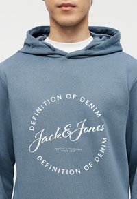 Κοντινό πλάνο ενός ατόμου που φοράει μπλε φούτερ με κουκούλα, με λευκό κείμενο «Jack & Jones» και «Definition of Denim» τυπωμένα σε κυκλικό σχέδιο.