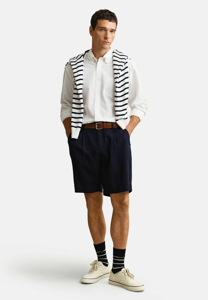 Man draagt een wit overhemd met knoopjes, marineblauwe korte broek, bruine riem, gestreepte trui over de schouders, zwart-wit gestreepte sokken en lichte sneakers.