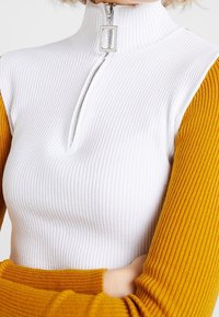 Ribbed turtleneck top featuring a white body and mustard long sleeves, with a partial zip closure and silver hardware accents.