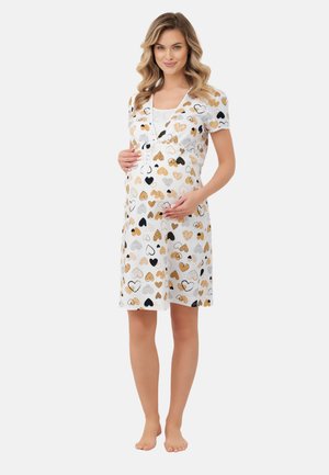 Kobieta w ciąży o falujących blond włosach, ubrana w białą sukienkę do kolan z czarnymi, beżowymi i brązowymi wzorami w kształcie serc, trzymająca się za brzuch.