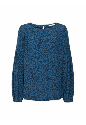 Nainen pukeutunut mustaan toppatakkiin, siniseen kuviolliseen paitaan, mustiin korkeavyötäröisiin kapealahkeisiin housuihin ja mustiin nilkkureihin, joissa on vetoketjut, seisoo valkoista taustaa vasten.