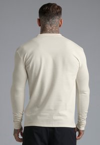 Muškarac s tetovažom na vratu, obučen u kremu majicu dugih rukava i crne hlače, okrenut leđima prema jednostavnoj sivoj pozadini.