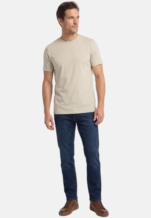 Mężczyzna stojący, ubrany w beżowy t-shirt z krótkim rękawem, ciemnoniebieskie dżinsy i brązowe skórzane trampki, patrzący w lewo.