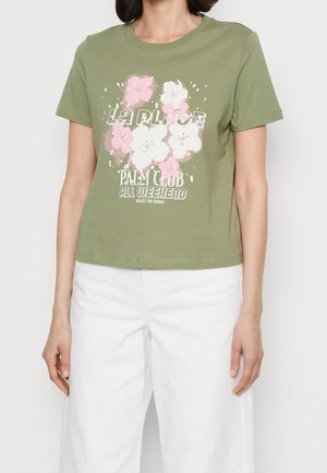 Mujer vistiendo una camiseta verde oliva con estampado floral en rosa y blanco y texto, combinada con pantalones blancos sobre un fondo liso.