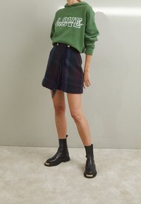 Groene hoodie met de tekst "LOVE", een geruite rok met franjes en zwarte enkellaarzen met een gouden accent op de zool. Over het geheel een gladde textuur.