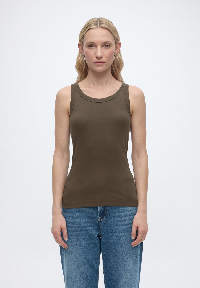 Ξανθιά γυναίκα που φοράει αμάνικη μπλούζα σε ελιά πράσινο και μπλε τζιν, στέκεται μπροστά σε απλό λευκό φόντο, κοιτάζοντας ευθεία μπροστά.