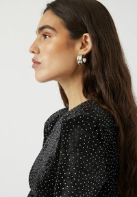 Pendientes de aro de plata con un acabado suave y brillante. Llevados por una persona con cabello largo y oscuro, combinados con una blusa de terciopelo negro con lunares.