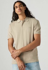 Beige polo skjorta med korta ärmar, krage och tre knappar. Slät textur med en diskret logotyp på vänster bröst.