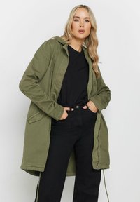 Giacca lunga con cappuccio in verde oliva, dotata di zip frontale, tasche laterali e vestibilità rilassata, abbinata a un top nero e pantaloni neri.