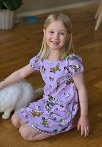 Violetti puuvillamekko lyhyillä hihoilla, jossa on värikkäitä sarjakuvahahmojen ja eläinten printssejä. Istuva lapsi valkoisen kissan kanssa lattialla.