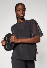 Zwart mesh sport t-shirt met korte mouwen en een ronde hals, voorzien van een subtiele logo. Model houdt een zwarte gewichtschijf vast.