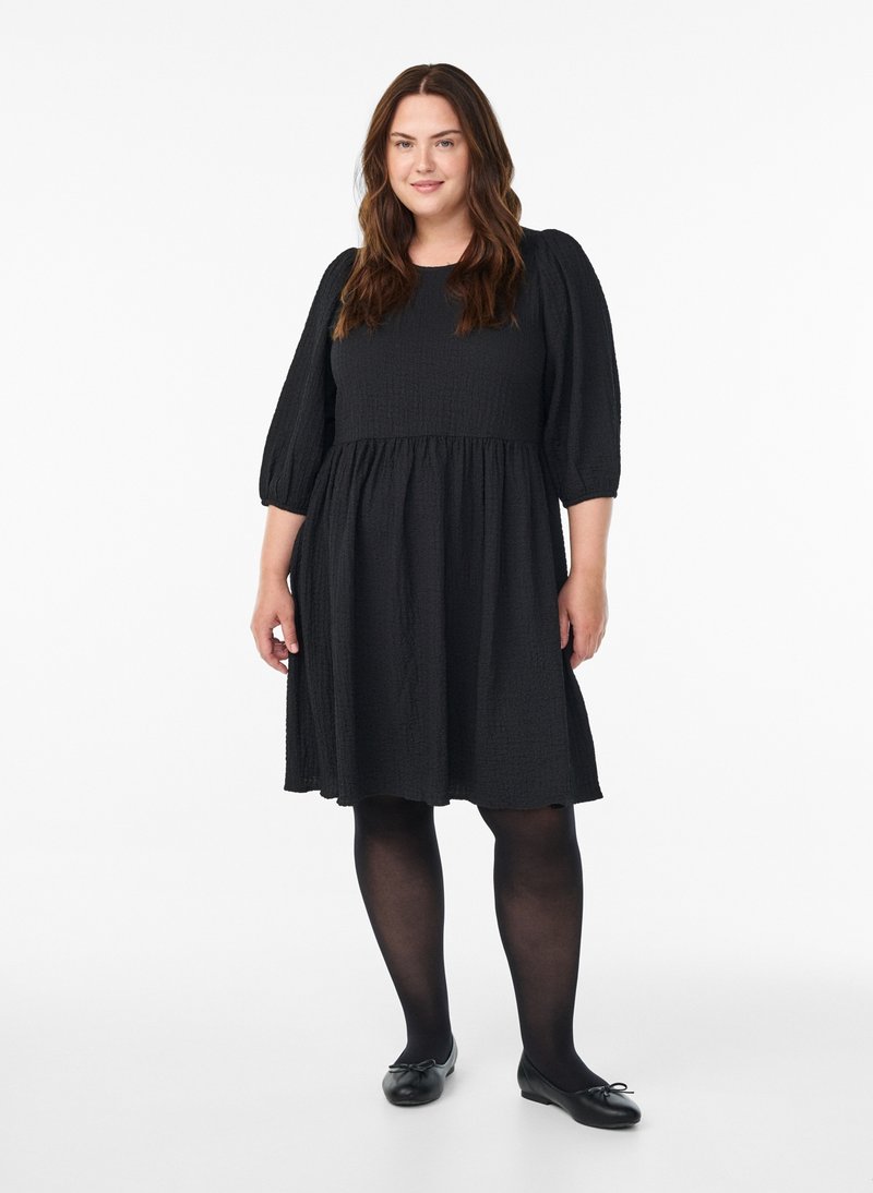 Mustast tekstuuriga puffy varrukatega kleit, põlveni pikk, liibuv ülakeha ja kogutud seelikuga. Kandatud koos mustade sukkpükste ja lamedate kingadega.