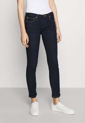 Personne portant un jean skinny bleu foncé et des baskets blanches, debout devant un fond clair uni.