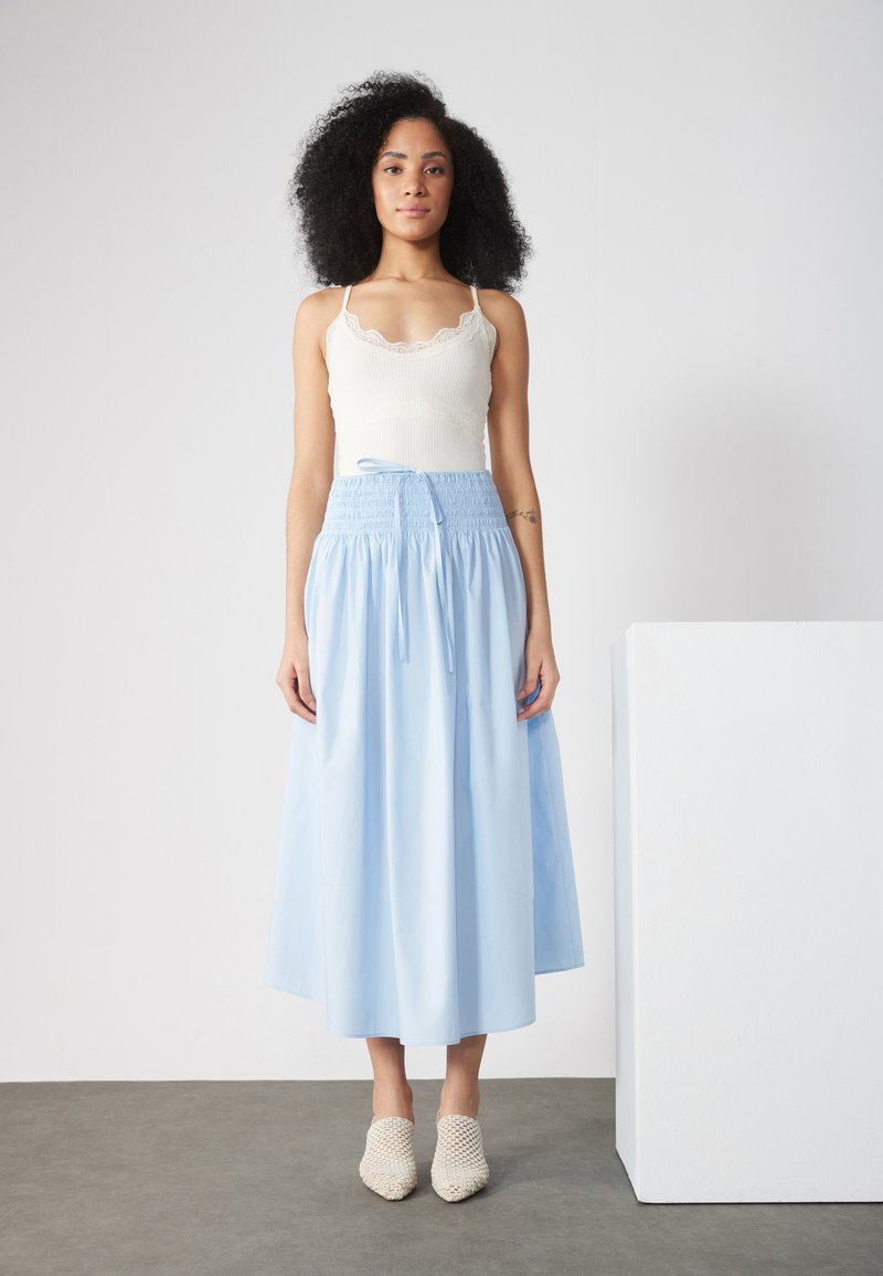 Jupe midi fluide bleu clair avec une taille smockée et un détail à nouer, associée à un débardeur blanc à dentelle et des chaussures en osier beiges.