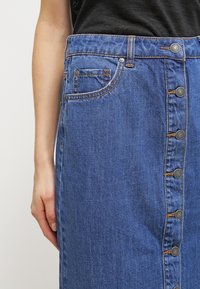 Denim-hame, jossa on sininen pesu, viisi nappia edessä ja sivutasku. Ompeleet vyötärön kohdalla.