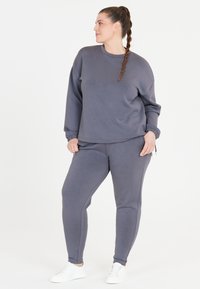 Grå sweatshirt och matchande byxor, tillverkade av mjukt tyg med en avslappnad passform. Byxorna har sidofickor och elastiska ärmslut. Vita sneakers.