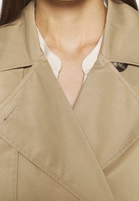 Béžový trenčkot s dvouvrstvým designem, s velkým knoflíkem a hladkou texturou s viditelnými švy.